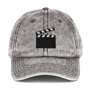 May include: A gray baseball cap with a distressed finish. The cap features a black embroidered film clapperboard design on the front. The clapperboard is open, showing the white stripes. The cap has a curved brim and an adjustable strap.
