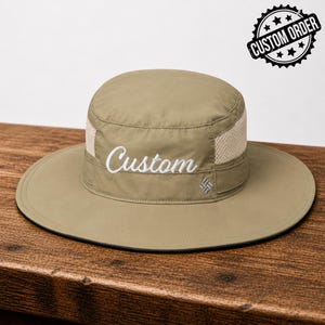 May include: A khaki-colored sun hat with a wide brim and mesh ventilation. The word "Custom" is embroidered in white on the front. A "Custom Order" stamp is visible in the upper right corner. The hat has a small logo on the side.
