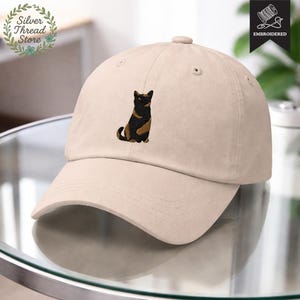 May include: A beige baseball cap with an embroidered design of a cat in black and brown. The cap has a curved brim and a button on top. The words "Silver Thread Store" are in a circular design on the left side of the cap.