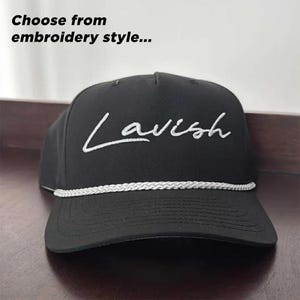 K&ouml;nnte beinhalten: Eine schwarze Baseballkappe mit dem in wei&szlig;er Schreibschrift auf das Vorderteil gestickten Wort Lavish. Eine wei&szlig;e, geflochtene Kordel verl&auml;uft horizontal &uuml;ber den unteren Teil der Kappe. Der Text Choose from embroidery style steht dar&uuml;ber.