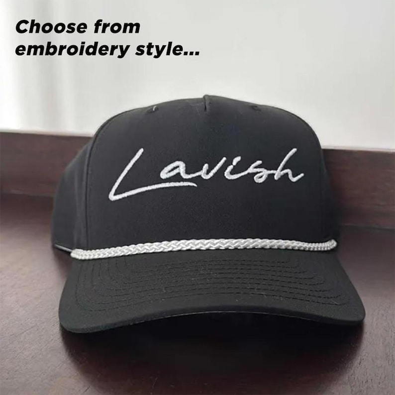 K&ouml;nnte beinhalten: Eine schwarze Baseballkappe mit dem in wei&szlig;er Schreibschrift auf das Vorderteil gestickten Wort Lavish. Eine wei&szlig;e, geflochtene Kordel verl&auml;uft horizontal &uuml;ber den unteren Teil der Kappe. Der Text Choose from embroidery style steht dar&uuml;ber.