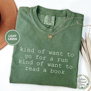 Op de afbeelding: Een saliegroen T-shirt met korte mouwen, een ronde hals en een Comfort Colors merklabel. Het shirt heeft witte tekst met de tekst: kind of want to go for a run kind of want to read a book. Een dunne gouden ketting ligt op de stof.