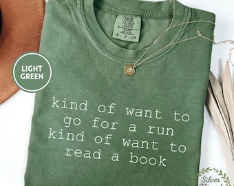 May include: A sage green short-sleeved t-shirt with a crew neckline and a Comfort Colors brand label. The shirt features white text that reads: kind of want to go for a run kind of want to read a book. A thin gold chain necklace rests on the fabric.