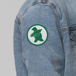 May include: Light blue denim jacket with a circular patch. The patch features a green bear holding a pencil on a white background with a green border. The jacket has a classic design with visible stitching and a buttoned front.