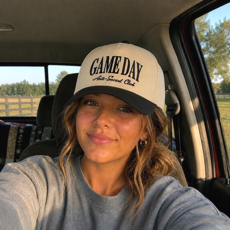 Op de afbeelding: Een beige en zwarte baseballcap met de tekst "GAME DAY Anti-Social Club" op de voorkant. De cap wordt gedragen door een persoon in een grijze trui, zittend in een voertuig.
