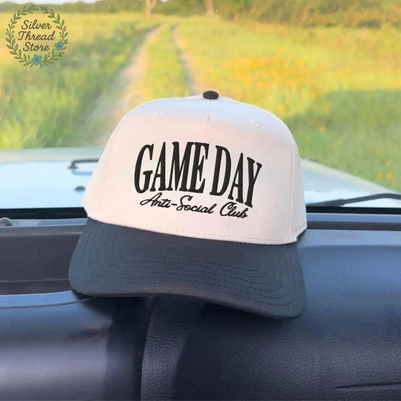 Op de afbeelding: Een wit en zwart baseball cap met de zwarte tekst "GAME DAY Anti-Social Club". De cap heeft een zwarte klep en een zwarte knop aan de bovenkant. De achtergrond toont een onverharde weg en groen.