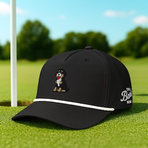 May include: Black baseball cap with a white rope detail and a small embroidered dog patch. The cap features the text "The Bark Nine" in white script on the side. The cap is on a green golf course.