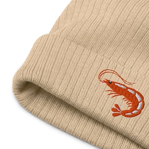 May include: A beige ribbed knit beanie featuring an embroidered orange shrimp design on the front. The shrimp has white accents and long antennae. The beanie is made of a soft, warm material.