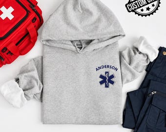 May include: A light gray hooded sweatshirt featuring the embroidered text ANDERSON above a dark blue Star of Life medical symbol. The hoodie is displayed alongside a red medical bag and dark navy blue cargo pants. A black CUSTOM ORDER stamp is visible.