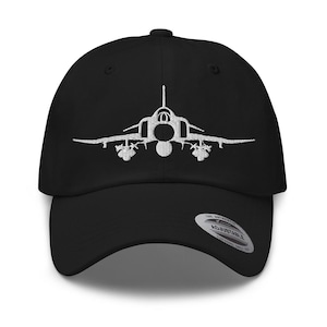 May include: Black baseball cap with a white embroidered image of a jet fighter plane. The cap has a curved brim and an adjustable strap at the back. The cap is made of a soft, durable fabric.
