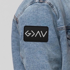 May include: Black embroidered patch with the text "G>AV" in white, attached to a denim jacket. The patch is rectangular with rounded corners. The jacket is a light blue wash.
