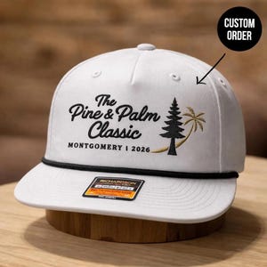 May include: A white five-panel cap featuring a black rope detail across the base of the crown. The front displays black embroidered text reading The Pine & Palm Classic MONTGOMERY | 2026, alongside a black pine tree and a gold palm tree icon.