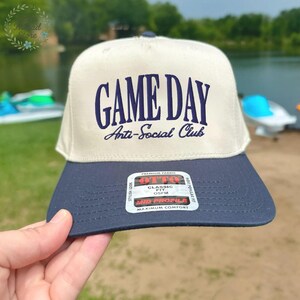Op de afbeelding: Een beige en marineblauwe baseballcap met de tekst "GAME DAY Anti-Social Club" in marineblauwe letters. De cap heeft een platte klep en een label met de tekst "OTTO Classic Fit OSFM Mid Profile Maximum Comfort".