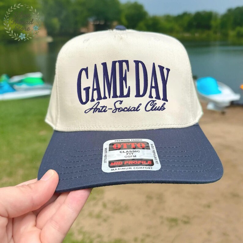 Op de afbeelding: Een beige en marineblauwe baseballcap met de tekst "GAME DAY Anti-Social Club" in marineblauwe letters. De cap heeft een platte klep en een label met de tekst "OTTO Classic Fit OSFM Mid Profile Maximum Comfort".