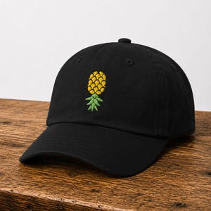 May include: Black baseball cap with a yellow and green embroidered pineapple design. The cap has a curved brim and an adjustable strap at the back. The pineapple is centered on the front of the cap.