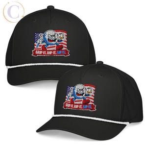 K&ouml;nnte beinhalten: Eine schwarze Baseballkappe mit einem wei&szlig;en Kordeldetail an der Basis der Krone. Die Vorderseite zeigt eine gestickte Grafik einer w&uuml;tenden Golfball-Figur mit einem Getr&auml;nk vor einer US-Flagge und dem Schriftzug GRIP IT. RIP IT. SIP IT.