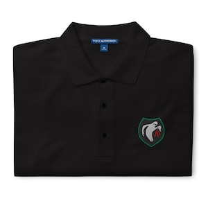 May include: Black polo shirt with a three-button placket and a small embroidered emblem on the chest. The emblem features a white ghost-like figure with red accents inside a green shield. The shirt is made by Port Authority.