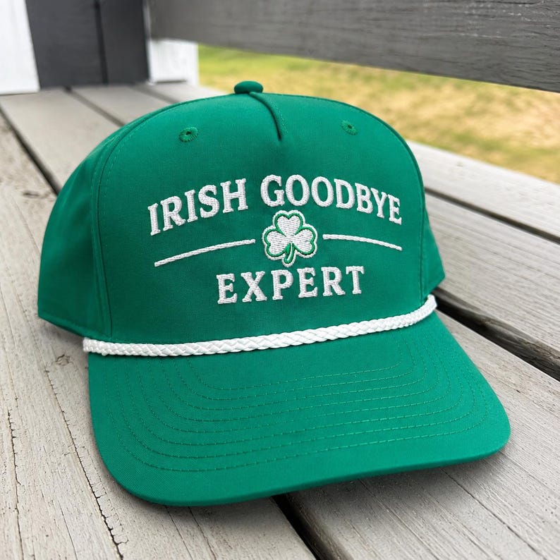K&ouml;nnte beinhalten: Eine leuchtend gr&uuml;ne Baseballkappe mit wei&szlig;em Sticktext, der IRISH GOODBYE EXPERT lautet, sowie einem kleinen wei&szlig;en Kleeblatt-Symbol in der Mitte. Die Kappe hat ein wei&szlig;es, geflochtenes Kordeldetail an der Basis des vorderen Bereichs.