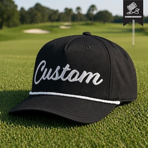 May include: Black baseball cap with white embroidered "Custom" script. A white rope detail trims the brim. The cap is on green grass. The word "Embroidered" is in the top right corner.