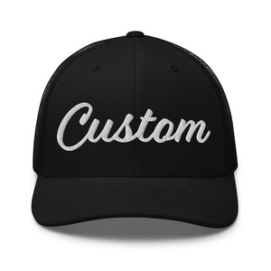 May include: Black trucker cap with a curved peak and mesh back. The word "Custom" is embroidered in white script across the front. A classic style of hat, suitable for casual wear. The hat is black.