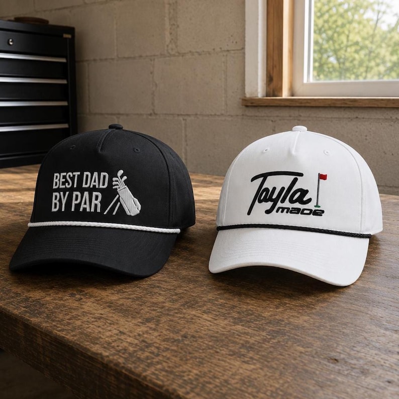 May include: A black baseball cap with white text reading BEST DAD BY PAR and a golf bag graphic, and a white baseball cap with black text reading Tayla made and a small red flag graphic. Both caps feature a decorative rope across the brim.