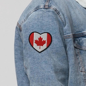 May include: A denim jacket with a heart-shaped patch featuring the Canadian flag. The patch has a red maple leaf on a white background, framed by red stripes and a black border. The jacket is light blue.
