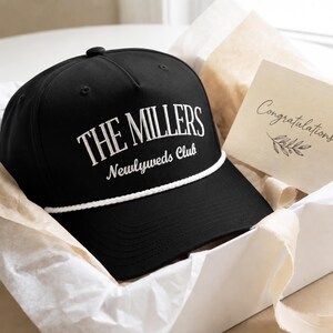 Op de afbeelding: Een zwarte honkbalpet met een wit koord over de klep en wit geborduurde tekst met de tekst THE MILLERS Newlyweds Club. Een klein kaartje met het woord Congratulations en een bladillustratie zit aan de zijkant van de doos gestoken.
