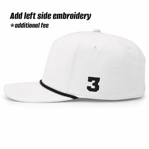 May include: A white baseball cap featuring a black braided cord across the base of the crown and a black embroidered number 3 on the side. The text Add left side embroidery *additional fee is visible at the top of the image.
