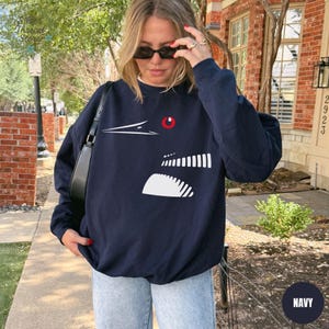May include: Navy blue sweatshirt featuring a white graphic design of a bird's head with a red eye. The sweatshirt has a crew neck and long sleeves. The word "NAVY" is in a white circle in the bottom right corner.