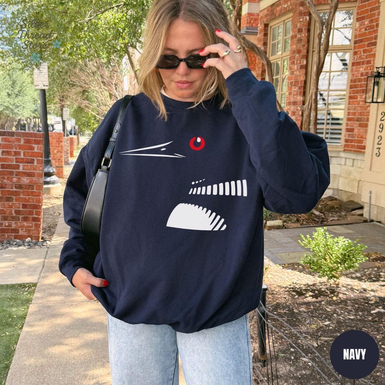 May include: Navy blue sweatshirt featuring a white graphic design of a bird's head with a red eye. The sweatshirt has a crew neck and long sleeves. The word "NAVY" is in a white circle in the bottom right corner.