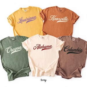 May include: Five short-sleeve crew neck t-shirts in mustard, yam, moss, ivory, and espresso colours. Each shirt features a location name printed in a cursive script font with an underline: Louisiana, Knoxville, Oregon, Alabama, and Columbia.