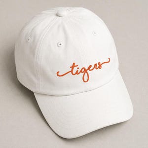 May include: A white baseball cap featuring the word tigers embroidered in orange cursive script across the front. The cap has a structured crown with a button on top, stitched eyelets for ventilation, and a curved brim.