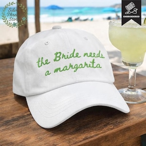 May include: White baseball cap with the embroidered phrase "the Bride needs a margarita" in green script. The hat is on a wooden surface, with a margarita cocktail in the background. The cap has a curved brim and a button on top.