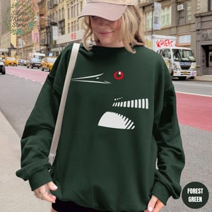 May include: Forest green sweatshirt featuring a white graphic design of a bird's head with a red eye. The sweatshirt has a crew neck and long sleeves. The words "Forest Green" are printed on a circular tag on the bottom right.