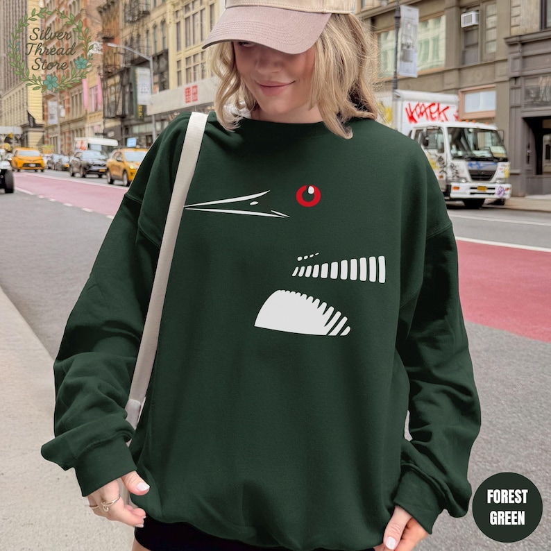 May include: Forest green sweatshirt featuring a white graphic design of a bird's head with a red eye. The sweatshirt has a crew neck and long sleeves. The words "Forest Green" are printed on a circular tag on the bottom right.