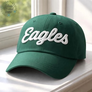 May include: A dark green baseball cap with the word "Eagles" embroidered in white script across the front. The cap has a curved brim and a button on top. The cap is sitting on a white surface.