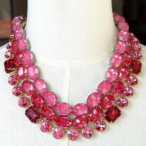 May include: A multi-strand pink gemstone necklace. The necklace features a variety of pink and red oval and round-cut gemstones. The gemstones are set in a gold-tone metal setting. The necklace is displayed on a white mannequin.