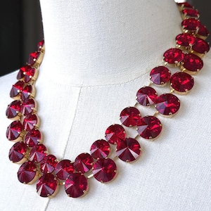 May include: A statement necklace featuring large, faceted, ruby-red crystals set in a gold-tone metal. The necklace is composed of multiple strands of the vibrant red stones, creating a bold and eye-catching design. The necklace is displayed on a white mannequin.