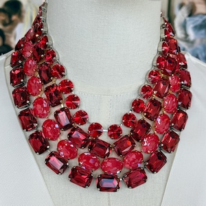 May include: A multi-layered statement necklace featuring numerous faceted red crystals in various shapes and sizes, including round and rectangular cuts. The vibrant red stones are set in a silver-toned metal frame, creating a dense, layered collar design.
