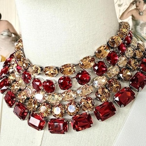May include: A multi-strand statement necklace featuring alternating rows of faceted red and champagne-colored crystals. The design consists of five layered strands of oval and rectangular cut stones set in silver-toned metal, creating a dense collar.