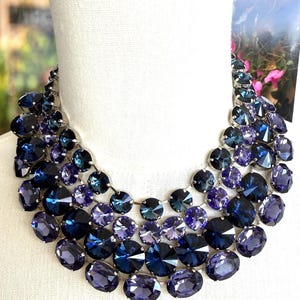 May include: A statement necklace featuring a cascade of large, faceted crystals in shades of blue and purple. The crystals are arranged in multiple rows, creating a bold and glamorous look. The necklace is displayed on a white mannequin.