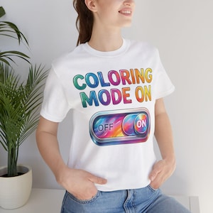 May include: White t-shirt with the text "COLORING MODE ON" in a rainbow gradient. Below the text is a colorful switch with "OFF" and "ON" labels. The shirt is a casual, everyday wear item.