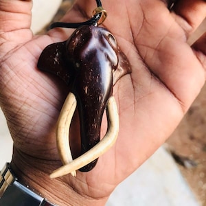 Może przedstawiać: Ciemnobrązowy wisiorek w kształcie słonia z kłami w kolorze kości słoniowej wisi na czarnym sznurku. Wisiorek jest trzymany w dłoni, ukazując misterne detale rzeźbionej głowy słonia. Unikalna biżuteria.