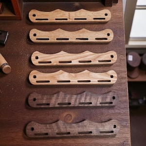 Op de afbeelding: Een verzameling van zes houten meshandvatten met een golvend ontwerp en drie sleuven. De handvatten zijn gemaakt van verschillende soorten hout, waaronder lichte en donkere bruintinten. Ze worden getoond op een houten oppervlak.