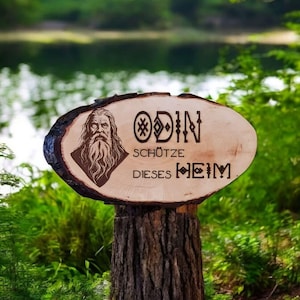 Könnte beinhalten: Ein Holzschild mit einer geschnitzten Abbildung von Odin, dem nordischen Gott, und dem Text "Odin schütze dieses Heim", was "Odin schütze dieses Heim" bedeutet.