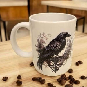 Può includere: Tazza in ceramica bianca con un'illustrazione dettagliata di un corvo nero posato su un design gotico ornato. Il corvo è raffigurato con intricati dettagli di piume. Chicchi di caffè sparsi intorno alla tazza.