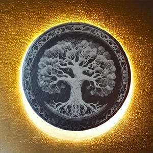 Op de afbeelding: Een ronde wandhanger van leisteen met een wit gegraveerd ontwerp van een levensboom op het oppervlak. De leisteen is omgeven door een decoratieve rand en wordt verlicht door warm wit licht.
