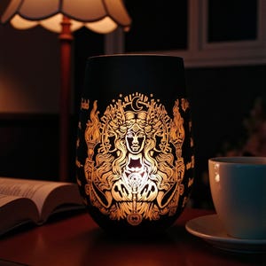 Könnte beinhalten: Schwarzer Glas-Kerzenhalter mit goldfarbenem Göttinnen-Design. Das Design zeigt eine zentrale Figur mit mehreren Gesichtern, Fackeln und einem Halbmond. Eine brennende Lampe und eine Tasse Kaffee befinden sich im Hintergrund.