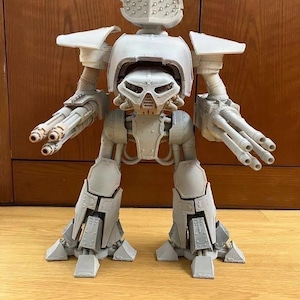 Könnte beinhalten: Ein hellgraues, unlackiertes Kunststoffmodell eines zweibeinigen Roboters mit einer großen, abgerundeten, am Kopf montierten Waffenanlage, einer zentralen schädelartigen Frontplatte und zwei mehrläufigen Kanonen an jedem Arm auf zwei Beinen.