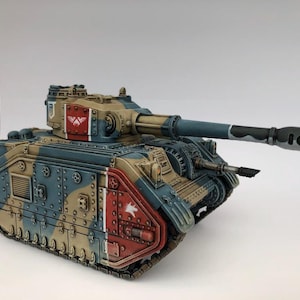 Puede incluir: Un modelo de tanque en miniatura pintado con un patrón de camuflaje de paneles beige, azules y rojos. El tanque tiene un cañón principal largo, un arma secundaria montada en la parte delantera, una antena fina e insignias blancas en la torreta.